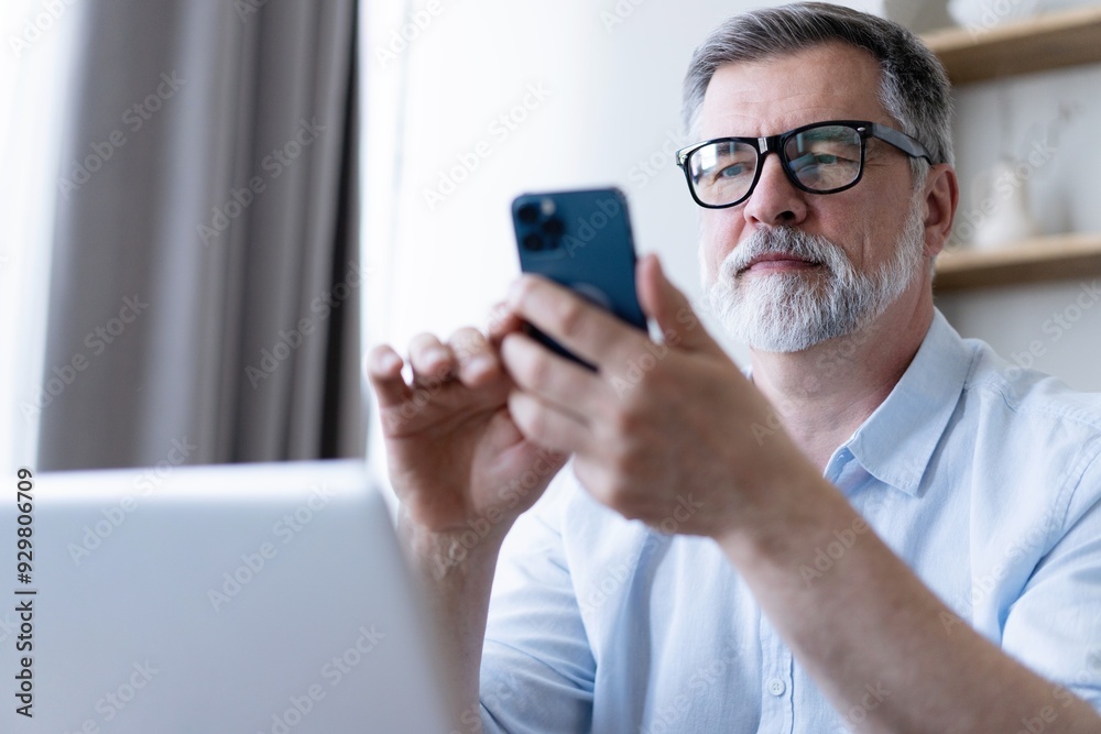 Handsome smiling senior man wearing glasses using mobile phone while sitting at his workplace with laptop at home