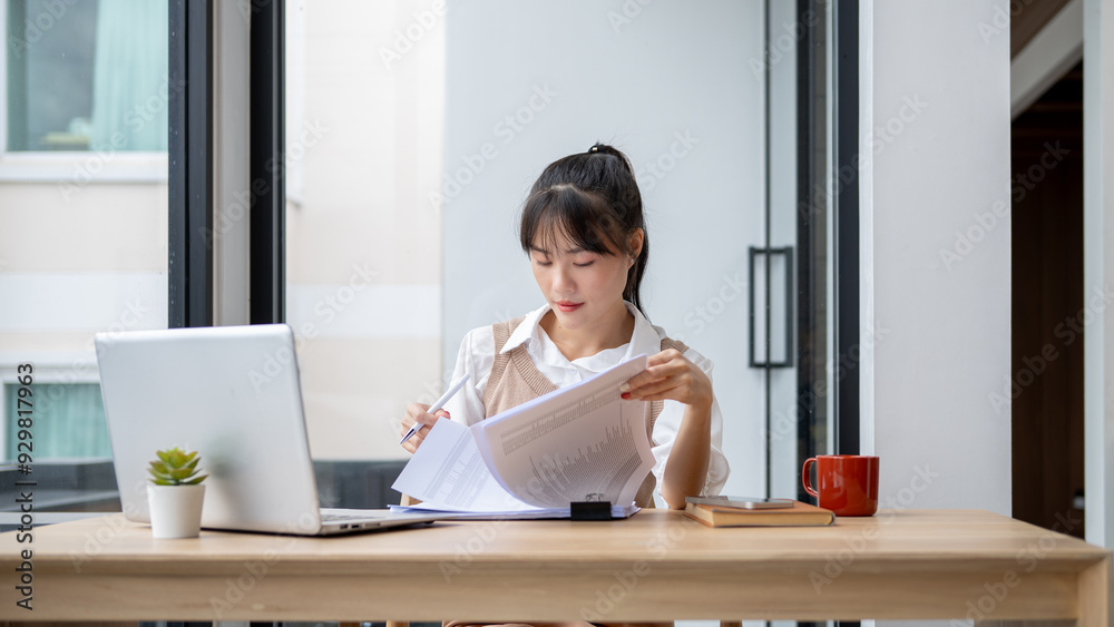 Obraz premium A focused Asian businesswoman is reviewing business report at her desk, working in her office.
