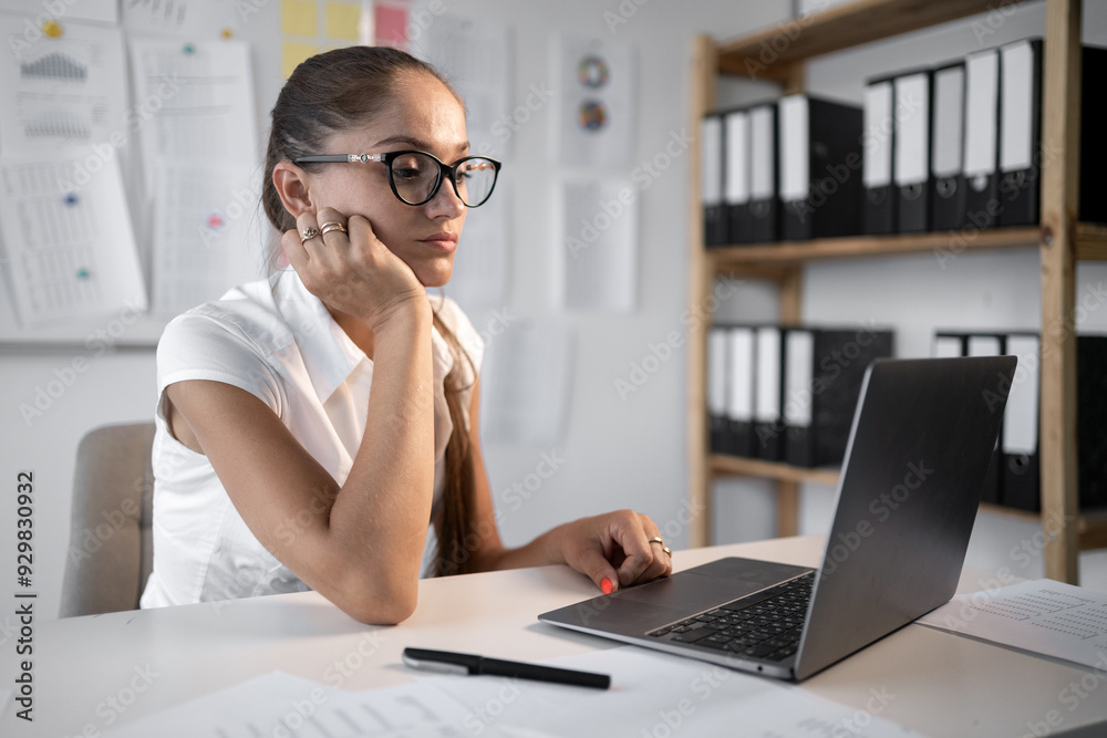 Confident businesswoman thought and watching laptop while sitting behind her computer and working in small office. Confident businesswoman thought and watching laptop while sitting behind her computer and working in small office.