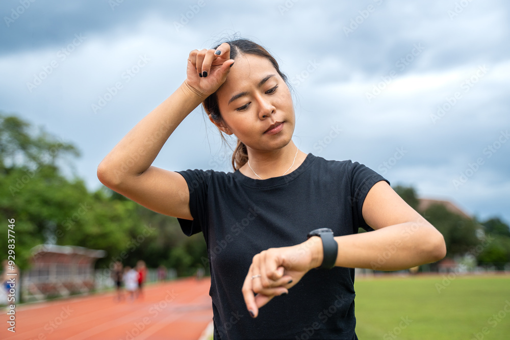 Fototapeta premium Young Asian female runner looking at her run time on her watch and wipe off her sweat from her forehead after her evening run