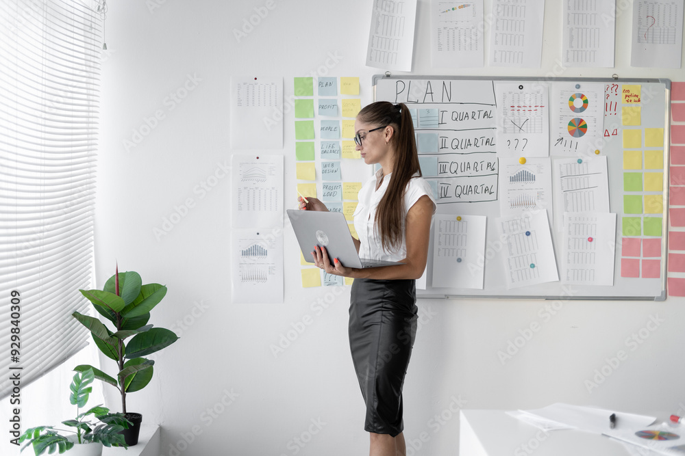 Concentrated young accountant with stickers near white board, preparing for meeting using laptop. Focused businesswoman explaining project ideas. Concentrated young accountant with stickers near white board, preparing for meeting using laptop. Focused businesswoman explaining project ideas.