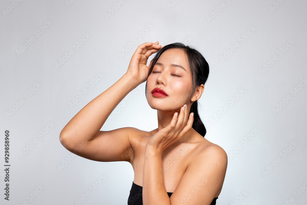 A beautiful Asian woman poses against a grey background, touching her face with her eyes closed. A beautiful Asian woman poses against a grey background, touching her face with her eyes closed.