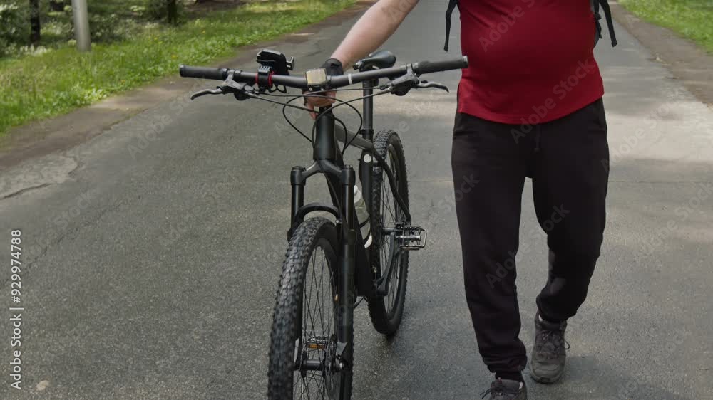 Senior man with gray beard wearing helmet walking with bicycle on road in woods and having talk on mobile phone Senior man with gray beard wearing helmet walking with bicycle on road in woods and having talk on mobile phone