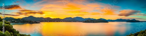 Panoramic view of a tranquil sunset over mountains and sea with orange clouds and blue water, serene landscape with green trees covering the land, creating a peaceful atmosphere in warm tones.