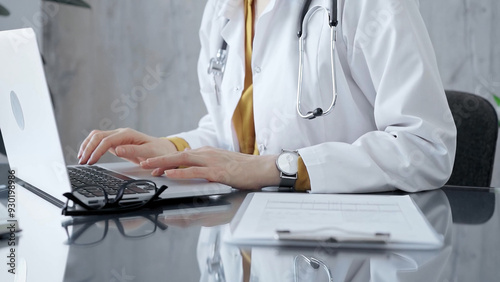 Doctor woman wearing white medical coat over a yellow blouse is working on laptop with stethoscope on the glass desk. Medicine and Health care