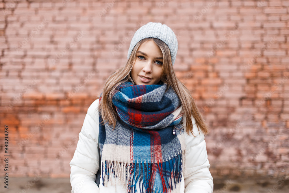 Happy beautiful young woman model in fashion clothes with knitted hat, scarf and winter jacket walking in the city near old brick wall Happy beautiful young woman model in fashion clothes with knitted hat, scarf and winter jacket walking in the city near old brick wall