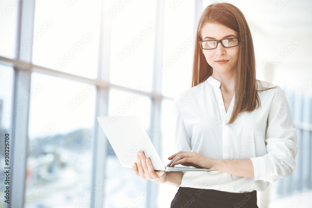 Beautiful female specialist with laptop computer standing in modern office and smiling charmingly. Working on design, data analysis, plan strategy. Business people concept Beautiful female specialist with laptop computer standing in modern office and smiling charmingly. Working on design, data analysis, plan strategy. Business people concept