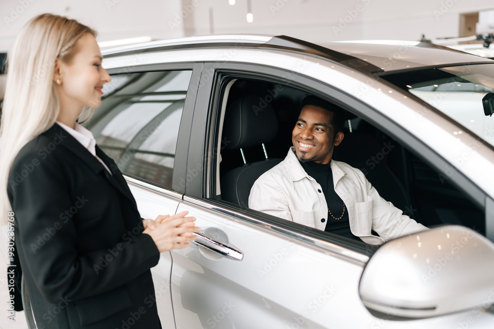 Cheerful African-American customer sitting behind steering wheel of new car in auto dealership, listening female consultant. Friendly auto dealer in business suit talking with black male customer. Cheerful African-American customer sitting behind steering wheel of new car in auto dealership, listening female consultant. Friendly auto dealer in business suit talking with black male customer.