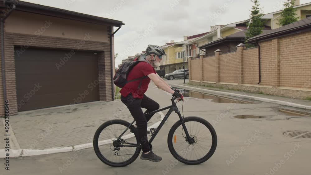 Side view tracking shot of senior man with backpack wearing helmet and sports clothes cycling along suburban street past residential buildings Side view tracking shot of senior man with backpack wearing helmet and sports clothes cycling along suburban street past residential buildings