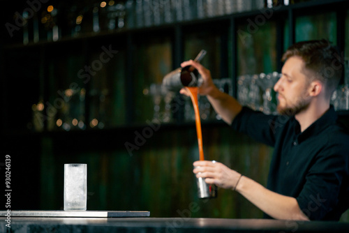 A young bartender in a black shirt prepares an alcoholic drink by pouring alcohol from  shaker in a shaker photo