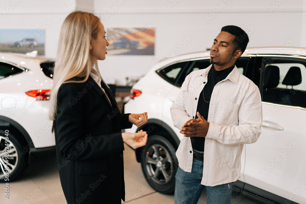 Side view of car saleswoman in business suit telling interested black male buyer about luxurious car in motor show. Serious African-American client looking at auto and listening to dealer. Side view of car saleswoman in business suit telling interested black male buyer about luxurious car in motor show. Serious African-American client looking at auto and listening to dealer.