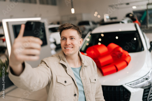 Closeup portrait of happy buyer male client doing selfie picture on smartphone at dealership after bought new car. Smiling young man choosing new vehicle in showroom and making photo using phone.