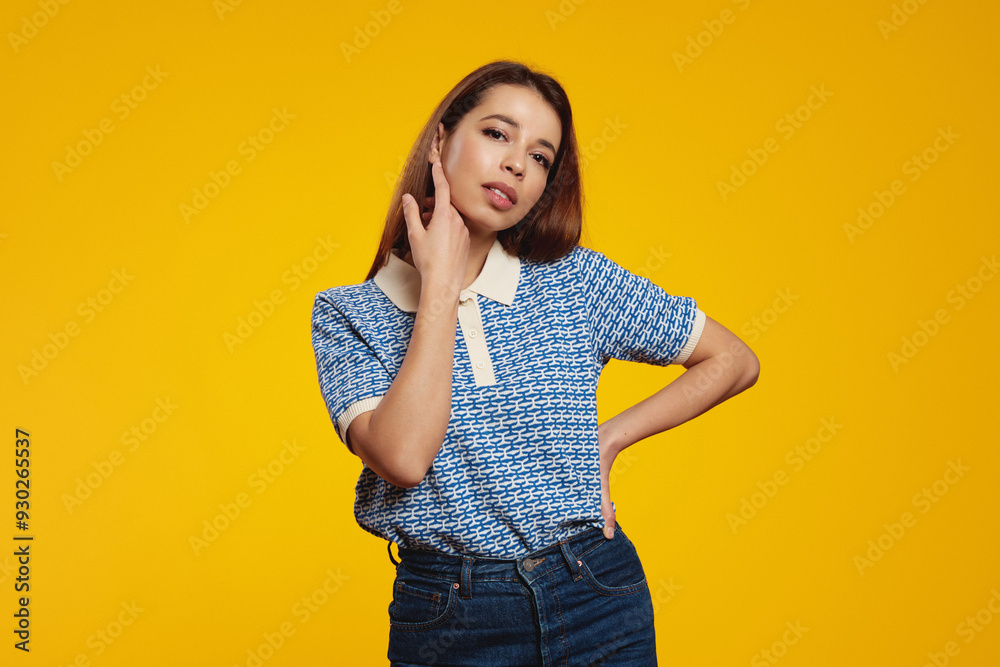 Attractive hispanic girl wearing blue shirt, touching her face with hand, standing and looking at camera against yellow background Attractive hispanic girl wearing blue shirt, touching her face with hand, standing and looking at camera against yellow background