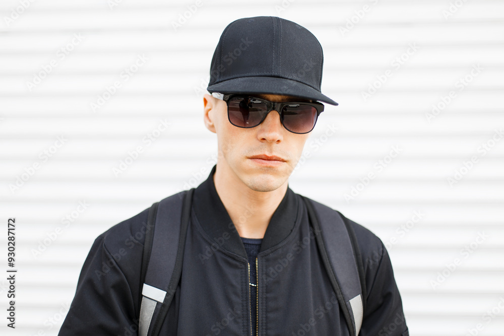 Cool fashion handsome young man model in black baseball cap with vintage sunglasses in black jacket with backpack on white metal background outdoors Cool fashion handsome young man model in black baseball cap with vintage sunglasses in black jacket with backpack on white metal background outdoors