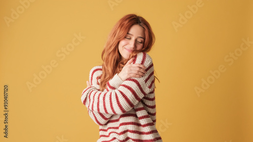 Cute red-haired woman dressed in sweater hugs herself looking at camera on yellow background. Beautiful girl poses in studio with blank space on yellow wall for your promotion