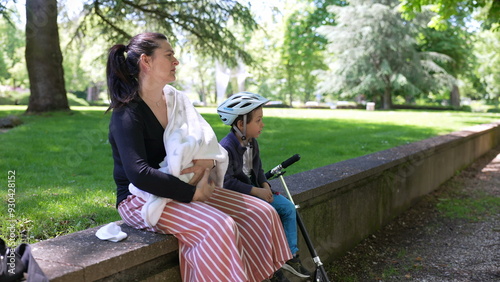 Mother breastfeeding baby on park bench, son wearing bike helmet sits beside with scooter, enjoying family time in nature, sunny day, peaceful park setting, maternal care and bonding