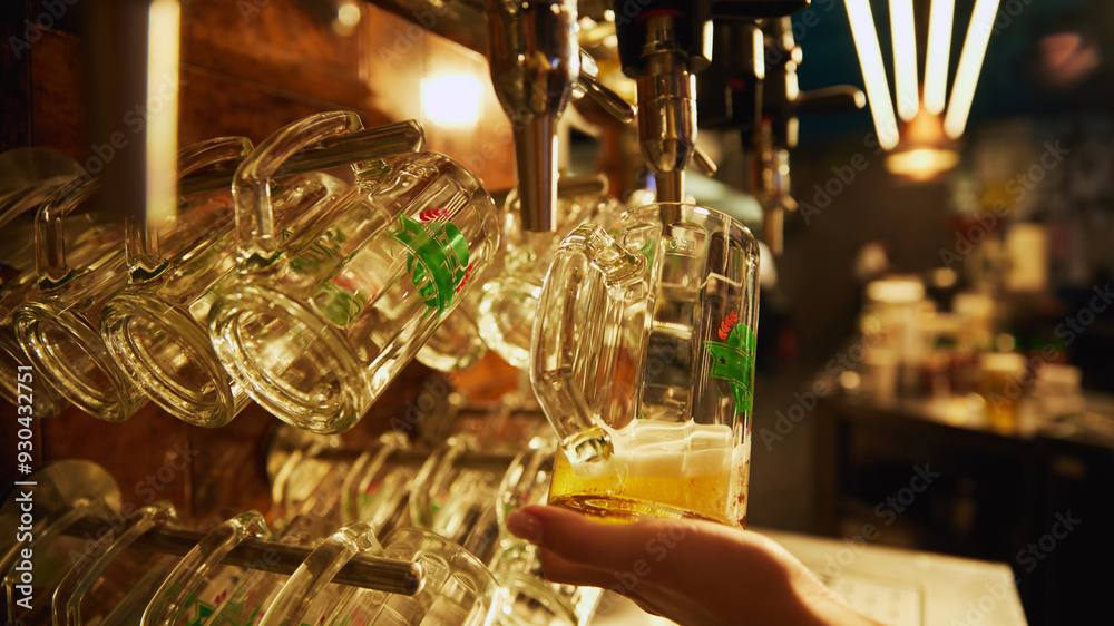 Bartender hand holding glass of lager beer being poured from tap, with rows of empty glasses hanging above and behind the tap. Concept of beer, brewery, pub, Oktoberfest, traditions Bartender hand holding glass of lager beer being poured from tap, with rows of empty glasses hanging above and behind the tap. Concept of beer, brewery, pub, Oktoberfest, traditions