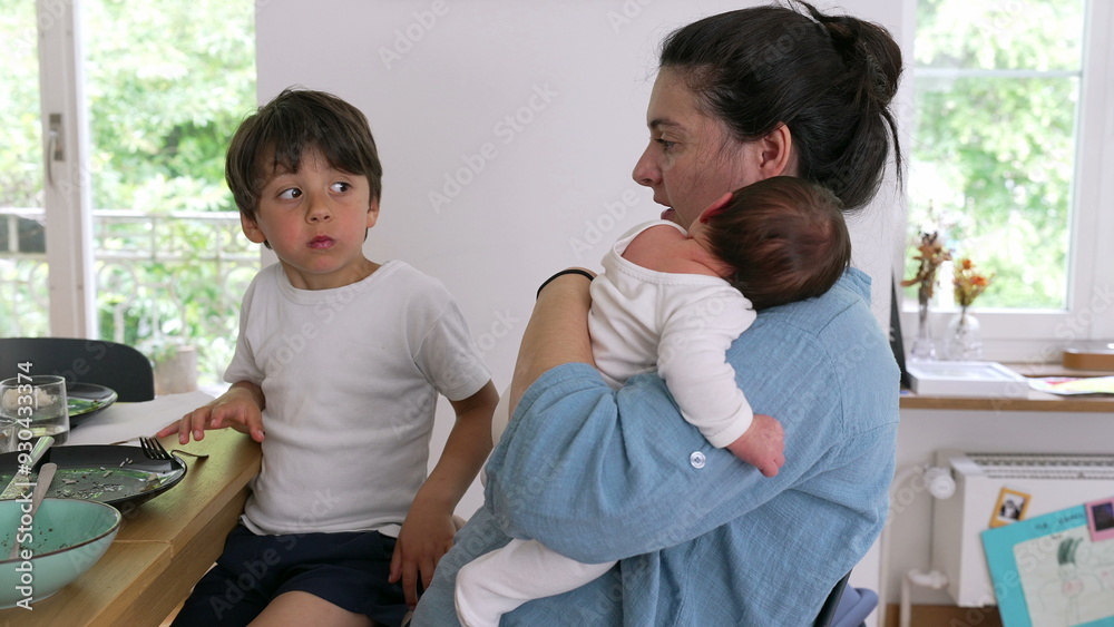 Mother holding baby while sitting next to an older child eating at the dining table. The scene reflects the multitasking and nurturing aspects of parenthood in a cozy home environment Mother holding baby while sitting next to an older child eating at the dining table. The scene reflects the multitasking and nurturing aspects of parenthood in a cozy home environment