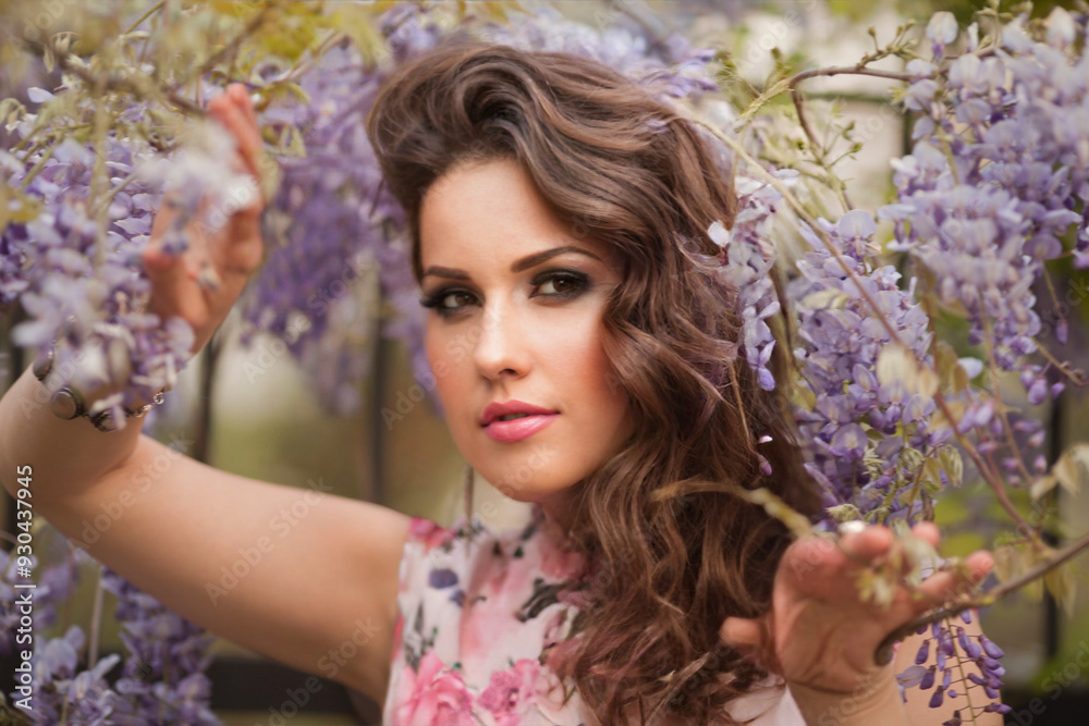 fashion spring outdoor portrait of beautiful young woman with long natural hair in luxurious dress posing among flowering wisteria trees in sunny garden