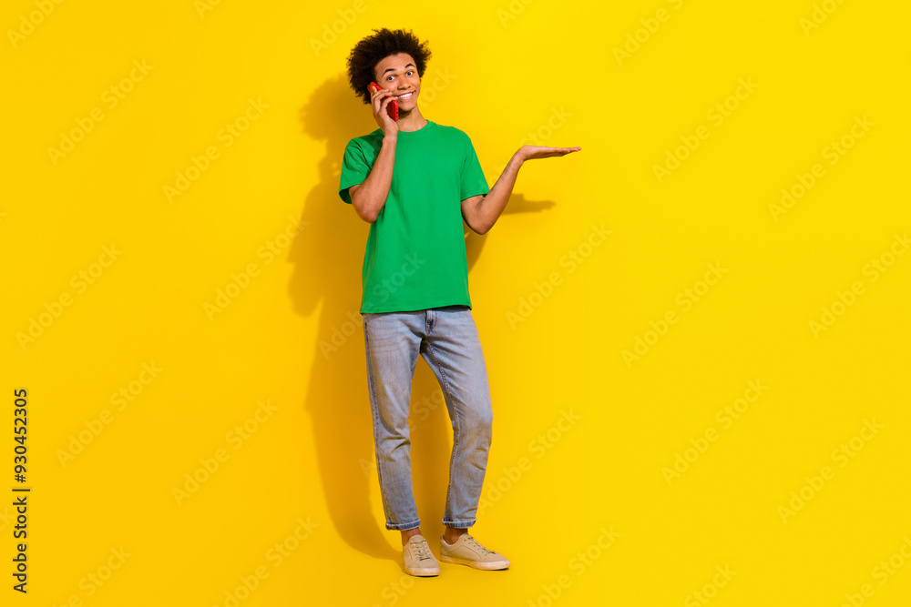 Full length photo of handsome good mood guy wear green t-shirt listening modern device showing arm emtpy space isolated yellow color background Full length photo of handsome good mood guy wear green t-shirt listening modern device showing arm emtpy space isolated yellow color background