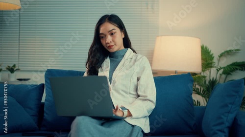 Young asian woman working remotely at night, sitting comfortably on a sofa while using a laptop. The cozy living room is softly illuminated by a lamp, creating a productive atmosphere