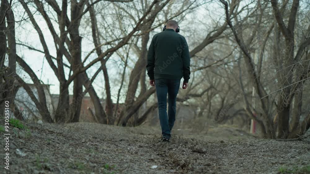 Man wearing a green jacket and headphones walking through a quiet forest, he is seen from behind, enjoying the peaceful surroundings of bare trees and a leaf-covered path Man wearing a green jacket and headphones walking through a quiet forest, he is seen from behind, enjoying the peaceful surroundings of bare trees and a leaf-covered path