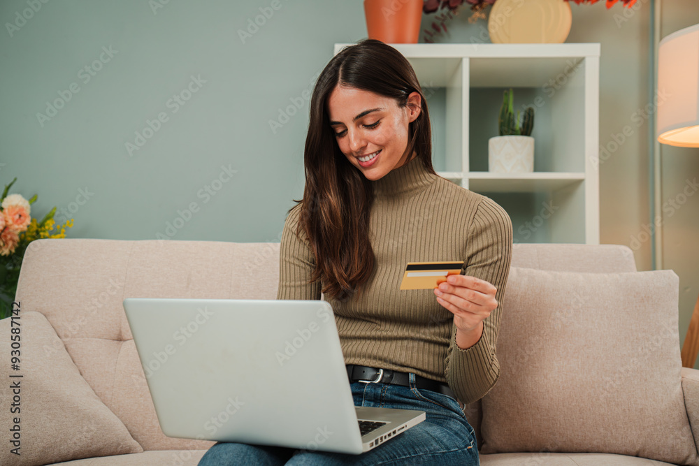 Young caucasian woman using a laptop to make an online purchase with a credit card sitting at home sofa. Female looking for a delivery with a computer at living room couch. Girl shopping online