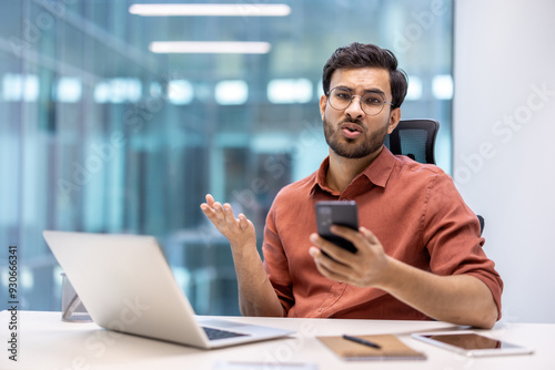 Man in office showing confusion holding smartphone, expressing frustration during work. He is sitting at desk with laptop and tablet, conveying business communication or technology issues.