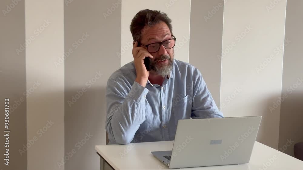 Smiling business man using laptop talking on cell phone sits at desk. Happy confident male professional manager web designer consulting client about online project making business call at workplace. Smiling business man using laptop talking on cell phone sits at desk. Happy confident male professional manager web designer consulting client about online project making business call at workplace.