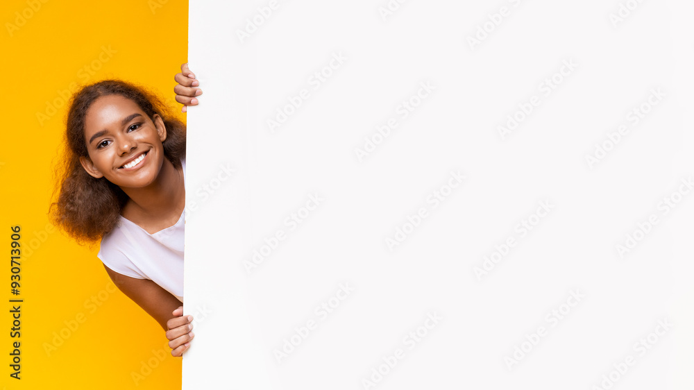 Joyful black girl looking from big white board for advertising, yellow background, panorama Joyful black girl looking from big white board for advertising, yellow background, panorama
