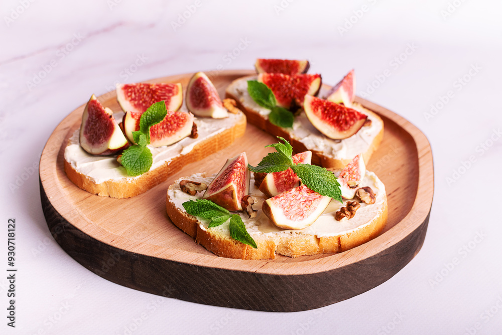Tasty homemade sandwiches with sweet figs, cream cheese, walnuts. honey and mint leaves on the wooden serving plate on light marble background close up.