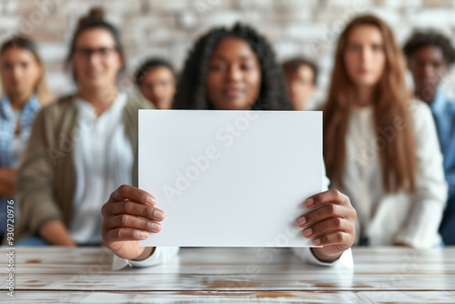A young African American woman holds a blank sheet of paper with space for messages and text, in the background a diverse group of coworkers sitting together, unity and collaboration