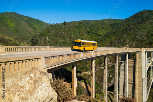A yellow school bus crossing a historic Bixby bridge surrounded by lush green hills under a clear blue sky