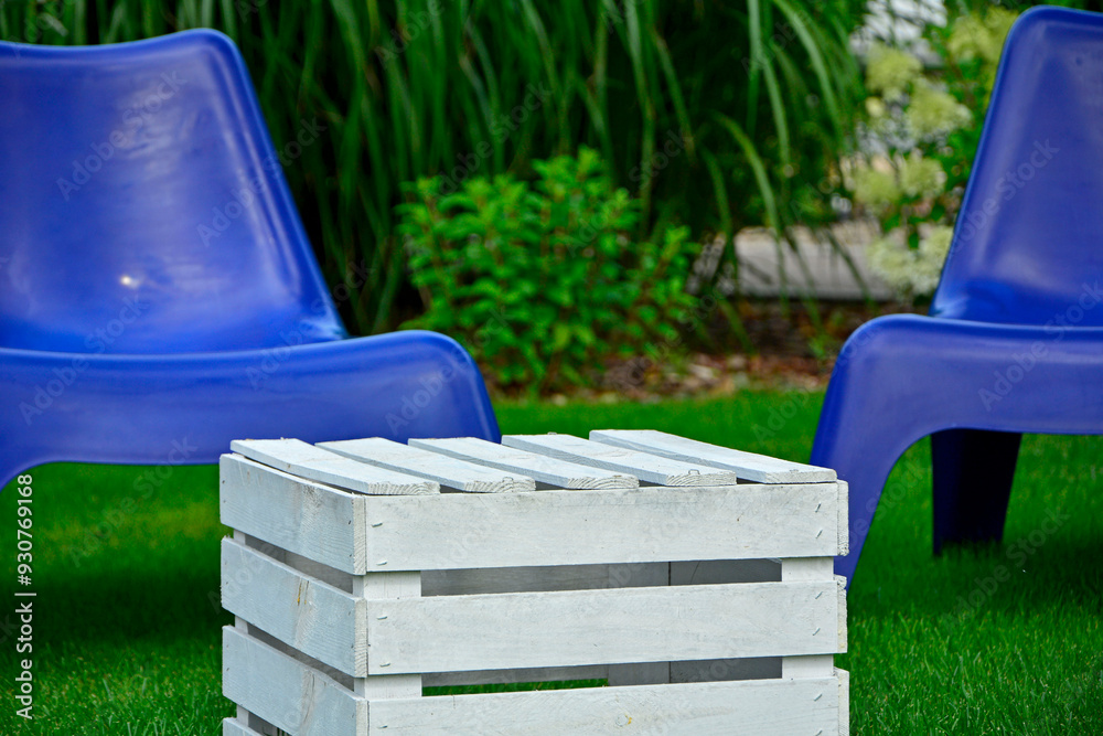biały stolik ze skrzynki i niebieskie fotele ogrodowe na trawie, blue plastic garden chair on grass, garden table made of box, blue chairs and white wooden box in garden, fotel ogrodowy