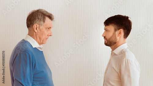Conflict Between Generations. Elderly Man And Mature Son Looking Severely At Each Other Standing Face-To-Face Against White Wall Indoor. Panorama