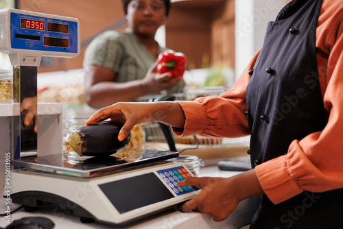 African american cashier weighing locally grown eggplant for female customer at the counter. Shopkeeper utilizing digital scale to measure weight of freshly harvested bio vegetable. photo