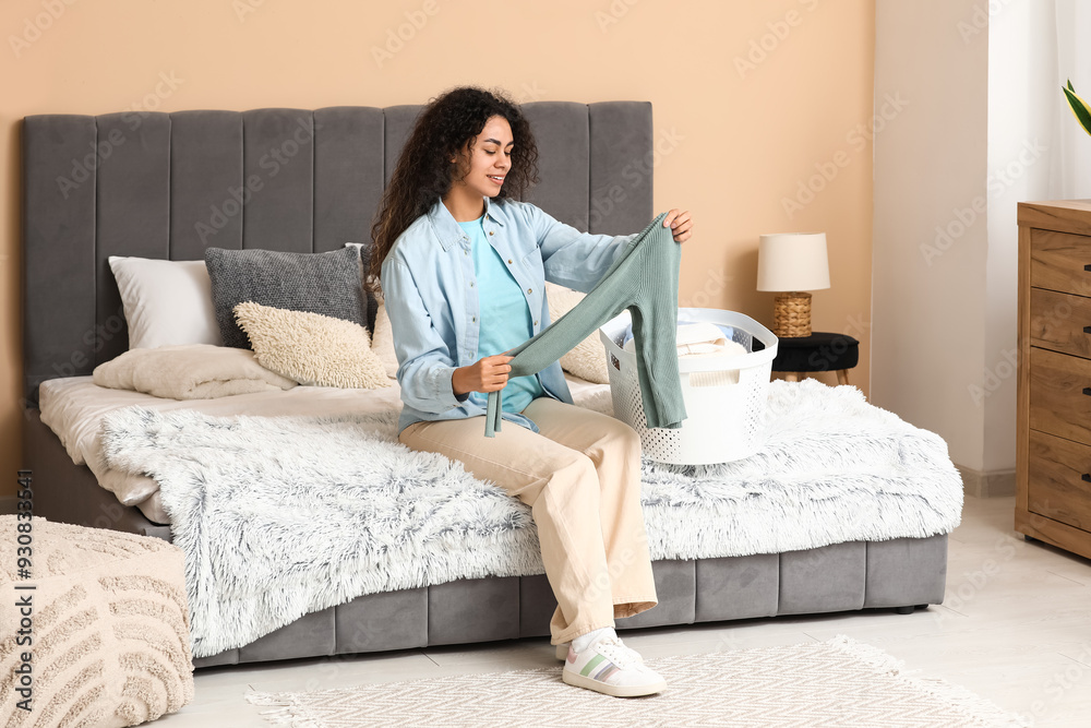 Beautiful young happy African-American woman with basket folding clean clothes in bedroom Beautiful young happy African-American woman with basket folding clean clothes in bedroom