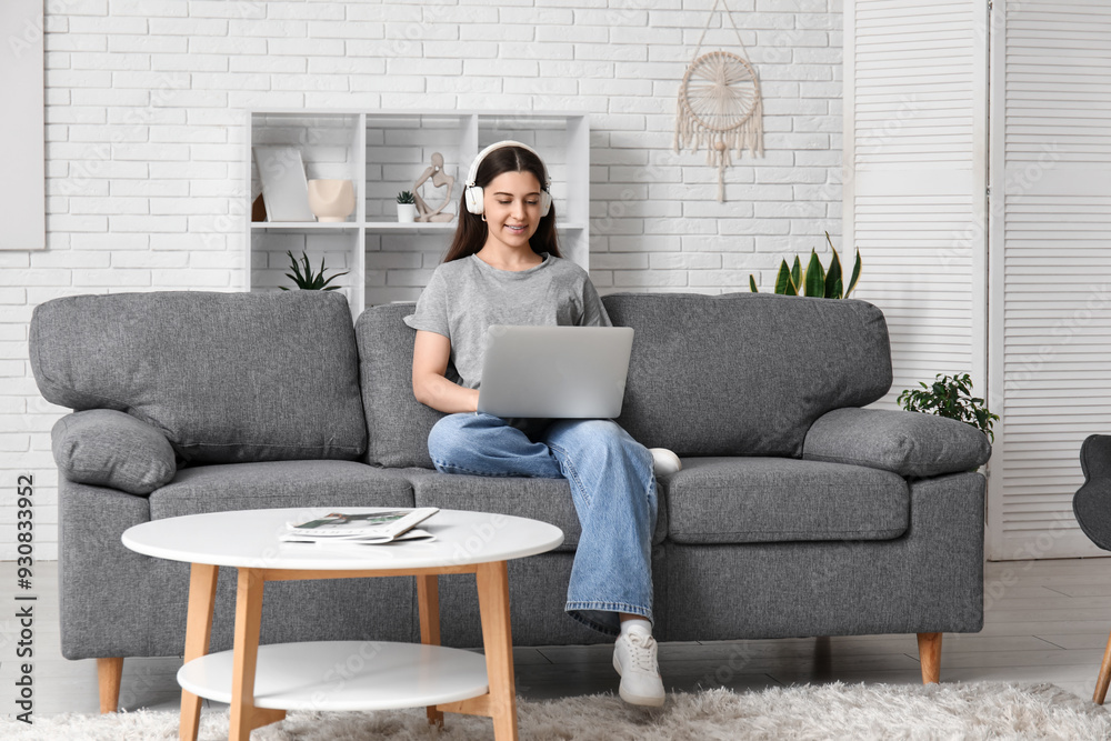 Young woman in headphones using laptop and sitting on black sofa in living room Young woman in headphones using laptop and sitting on black sofa in living room