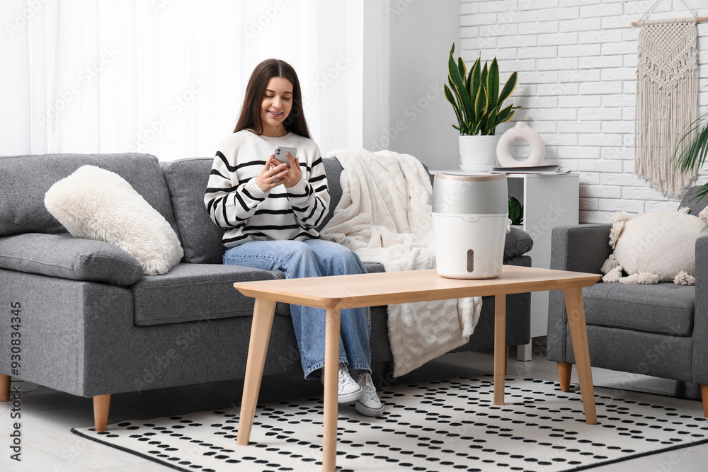 Young woman chatting with mobile phone on sofa near modern humidifier in living room Young woman chatting with mobile phone on sofa near modern humidifier in living room