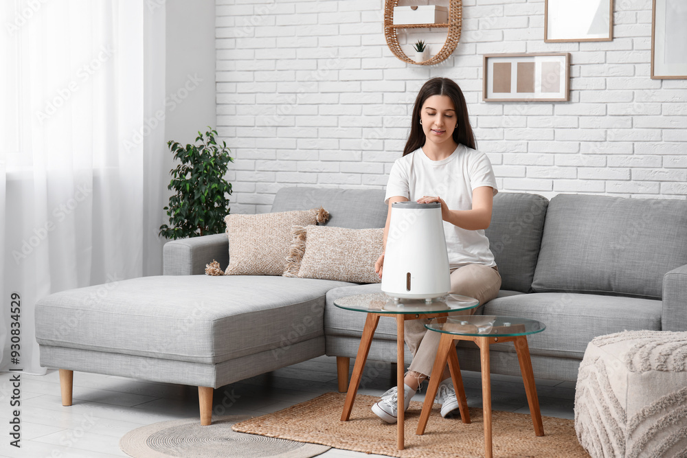 Young woman sitting on cozy sofa and turning on modern humidifier in stylish living room Young woman sitting on cozy sofa and turning on modern humidifier in stylish living room