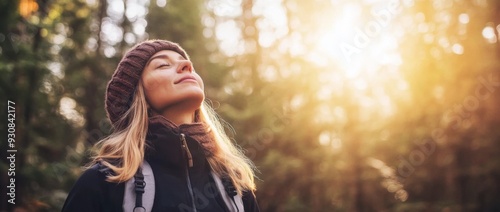 Woman in winter clothing stands in the forest with her eyes closed, breathing deeply and enjoying sunlight filtering through trees. Peaceful and happy expression with a warm golden glow.