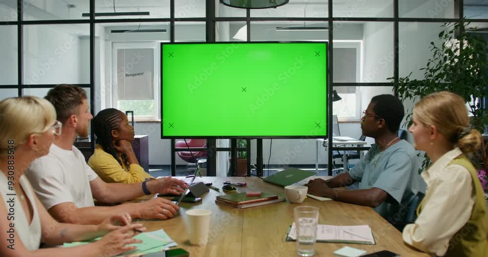 A group of young people are looking at a green screen while sitting at a table in an office meeting room. Copy space, mockup