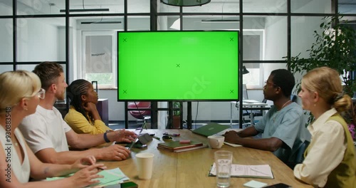 A group of young people are looking at a green screen while sitting at a table in an office meeting room. Copy space, mockup