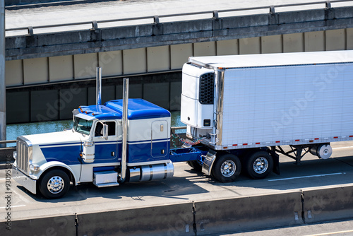 Blue and white classic big rig semi truck with high exhaust pipes transporting cargo in refrigerated semi trailer running on the overpass road intersection along the river photo