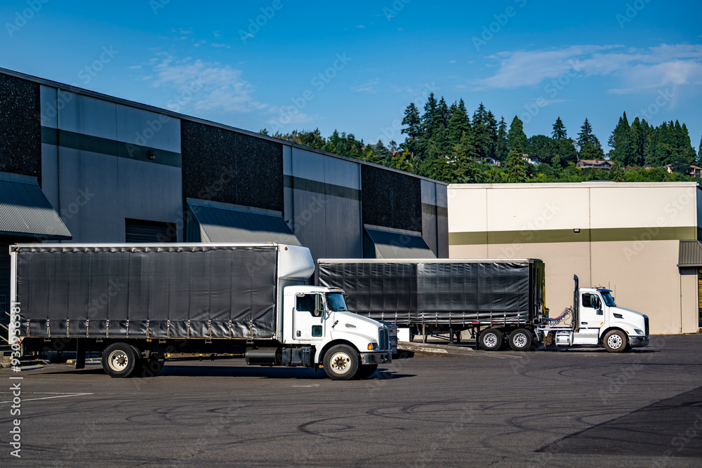 Day cab white big rig semi trucks with Conestoga trailers loading cargo standing in warehouse dock gates Day cab white big rig semi trucks with Conestoga trailers loading cargo standing in warehouse dock gates