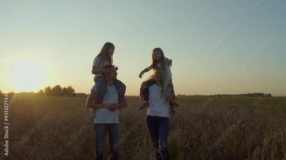 Carefree young couple walks through wheat field at sunset with their daughters. girls are sitting on their parents' shoulders and laughing. Family vacation in nature Carefree young couple walks through wheat field at sunset with their daughters. girls are sitting on their parents' shoulders and laughing. Family vacation in nature