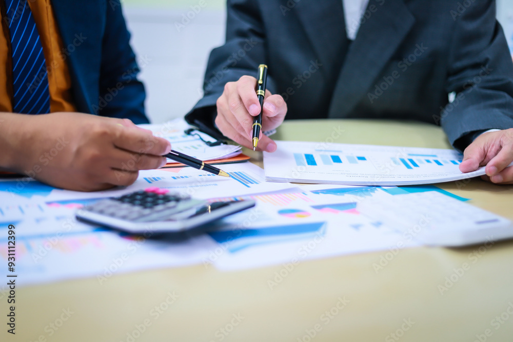 Two businessmen are seated at a desk in an office, engaged in a meeting focused on analysis, finance, and marketing strategies, with documents and graphs spread across the table. Two businessmen are seated at a desk in an office, engaged in a meeting focused on analysis, finance, and marketing strategies, with documents and graphs spread across the table.