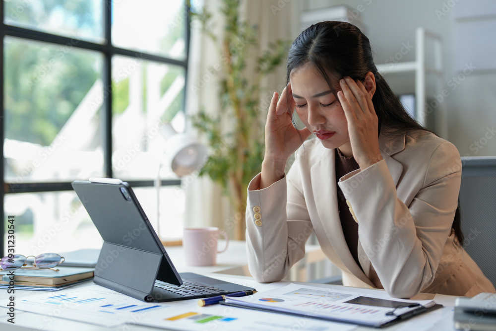 Tired Asian woman working with tax documents on laptop computer in office. Sad, worried, depressed or stressed employee life. Stretch your muscles due to sitting and working for a long time. Tired Asian woman working with tax documents on laptop computer in office. Sad, worried, depressed or stressed employee life. Stretch your muscles due to sitting and working for a long time.