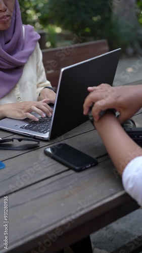 A Muslim woman in a hijab works on her laptop at an outdoor wooden table while a colleague helps plug in a cable, showcasing modern collaborative remote work and connectivity.