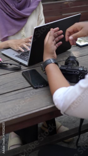 Diverse professionals collaborating on a creative project at an outdoor workspace, discussing ideas over a laptop with a camera and smartphone on the table.